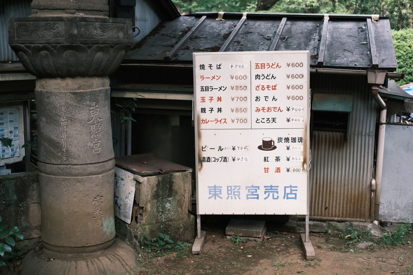 Signboard displaying menu outside a traditional Japanese food stall, surrounded by greenery.
