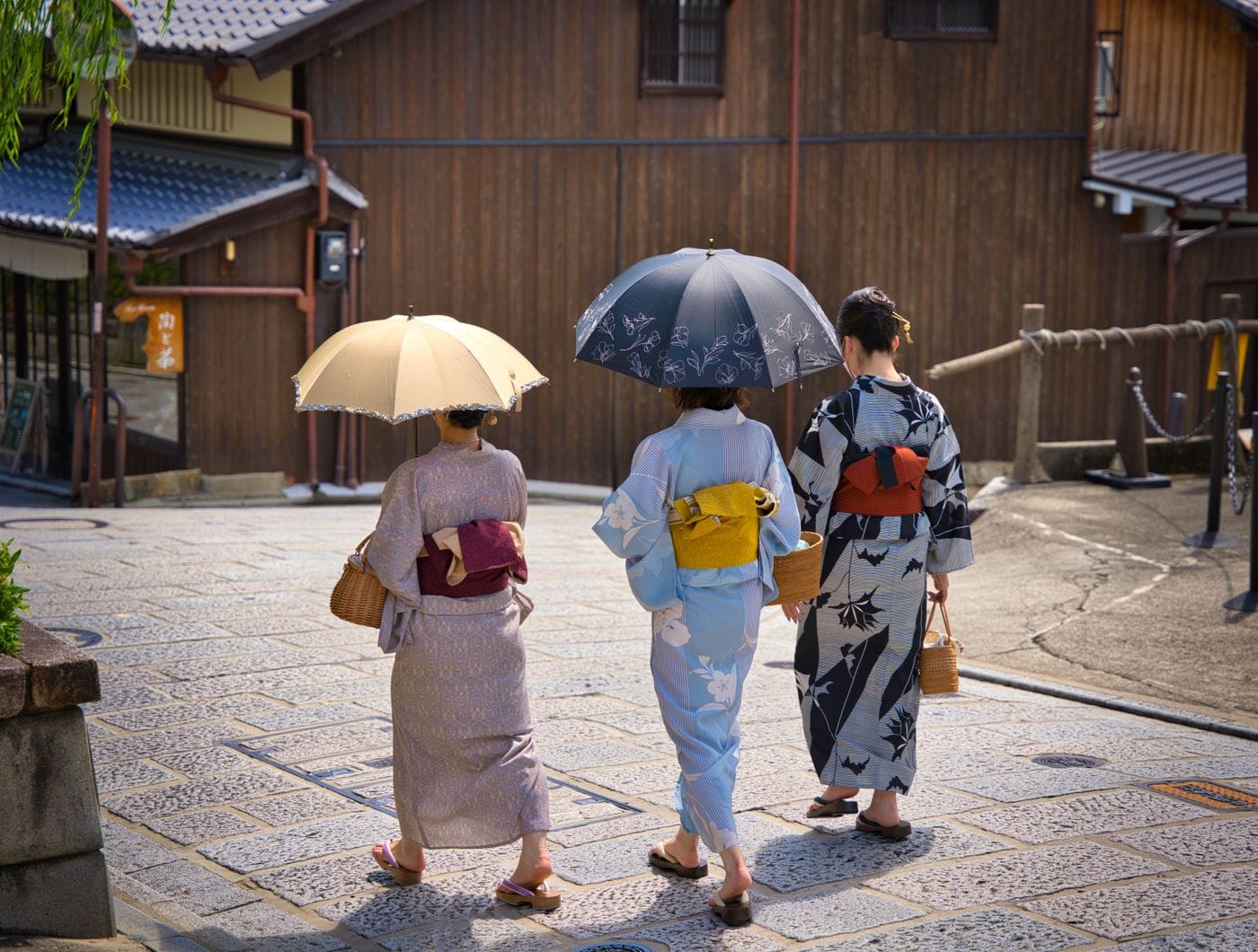 Three women in kimono with umbrellas strolling through Kyoto's traditional streets, showcasing cultural elegance.