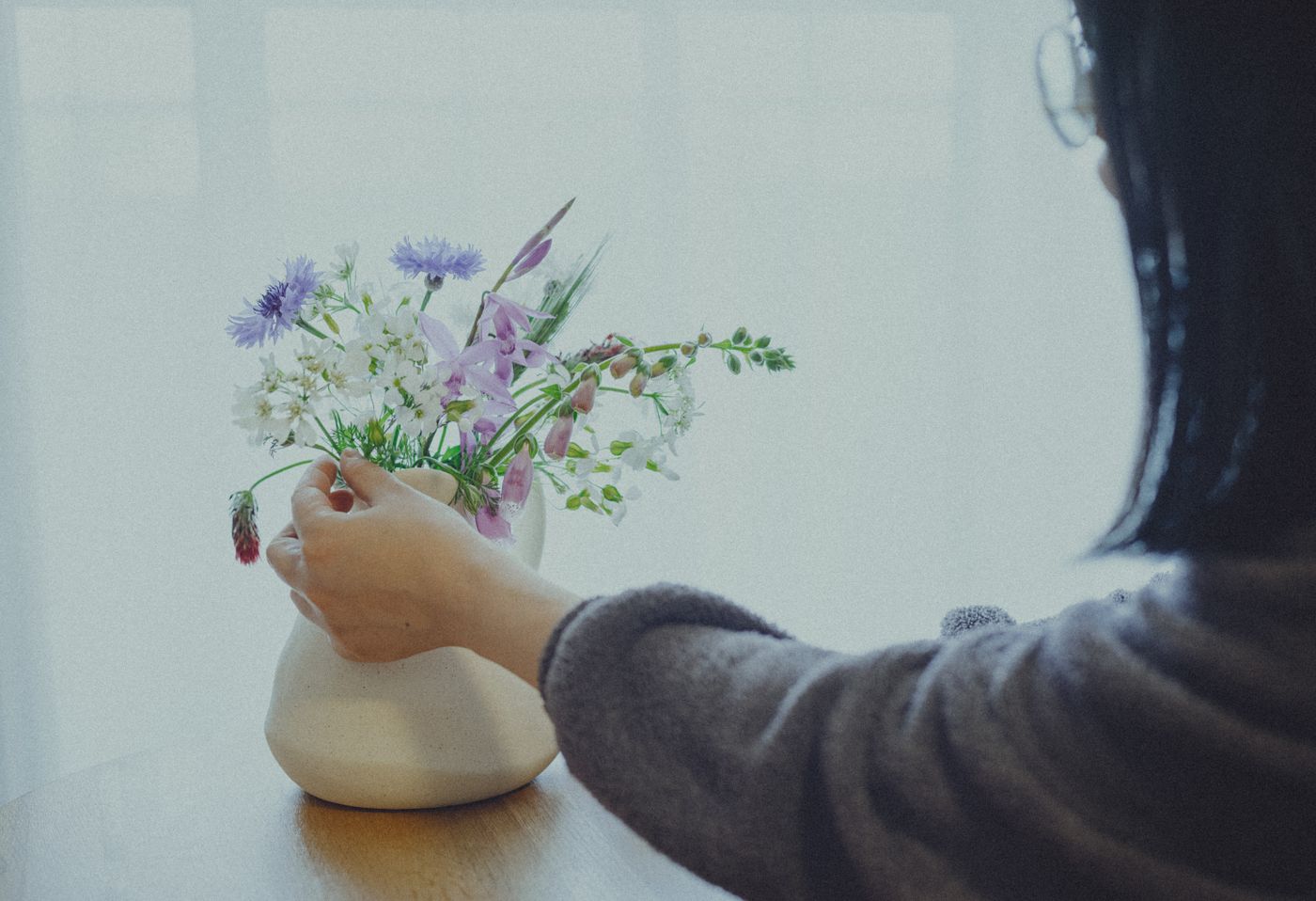 A person arranges a beautiful ikebana flower arrangement in a white vase indoors, showcasing traditional Japanese floral art.
