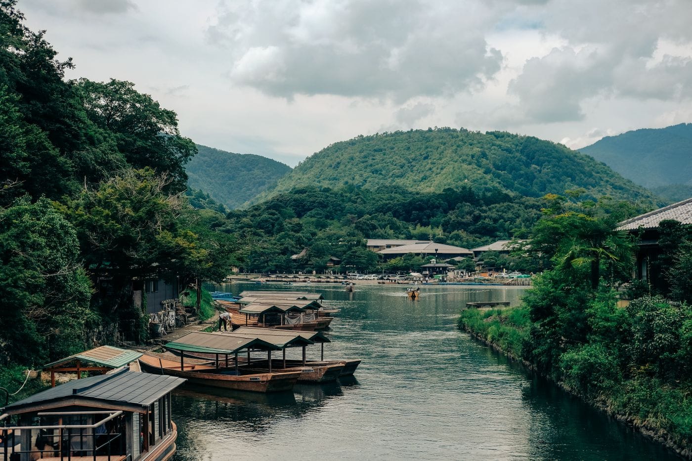 Tranquil riverside scene with wooden boats in Arashiyama, Kyoto, surrounded by lush green mountains.