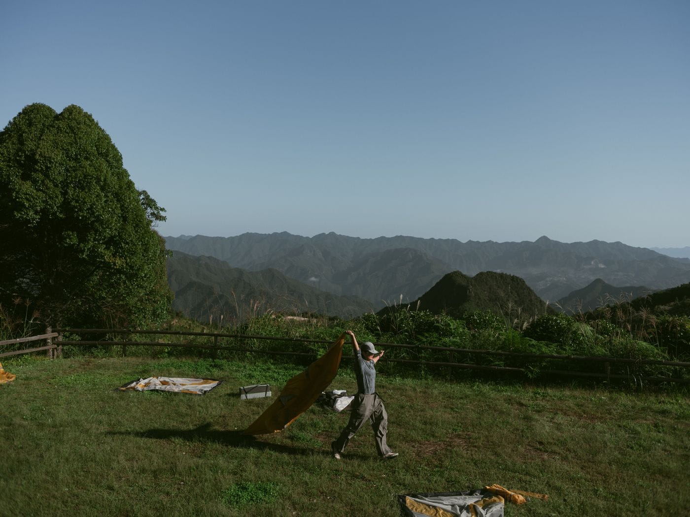 An individual prepares a tent on a grassy field against a backdrop of mountains under a clear sky.