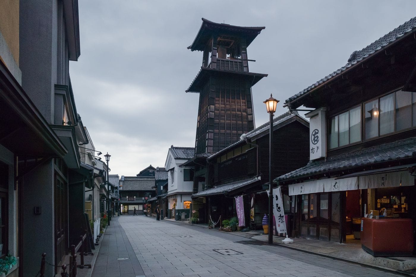 A quiet street in Japan featuring a traditional wooden bell tower and vintage architecture under a cloudy sky.